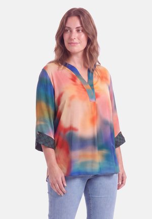 Vrouw draagt een losse blouse met lange mouwen met een kleurrijk tie-dye patroon en lichtblauwe spijkerbroek, staand tegen een effen achtergrond.