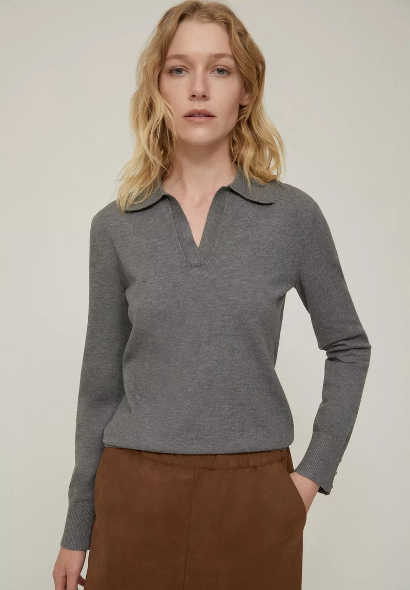 Pullover grigio a maniche lunghe con scollatura a V e colletto, realizzato in materiale morbido. Abbinato a una gonna marrone, mani in tasca.