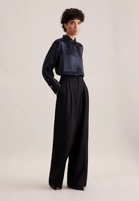Black satin long-sleeve shirt with a pointed collar, paired with high-waisted, wide-leg black trousers and pointed black heels.
