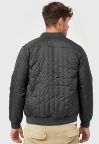 Padded black bomber jacket with vertical quilted texture, ribbed cuffs, and hem. Back view shows a simple collar and streamlined design.