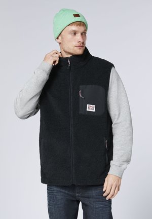 Man wearing light green beanie, gray sweatshirt, black fleece vest with zip-up and chest pocket, adjusting collar while looking right.