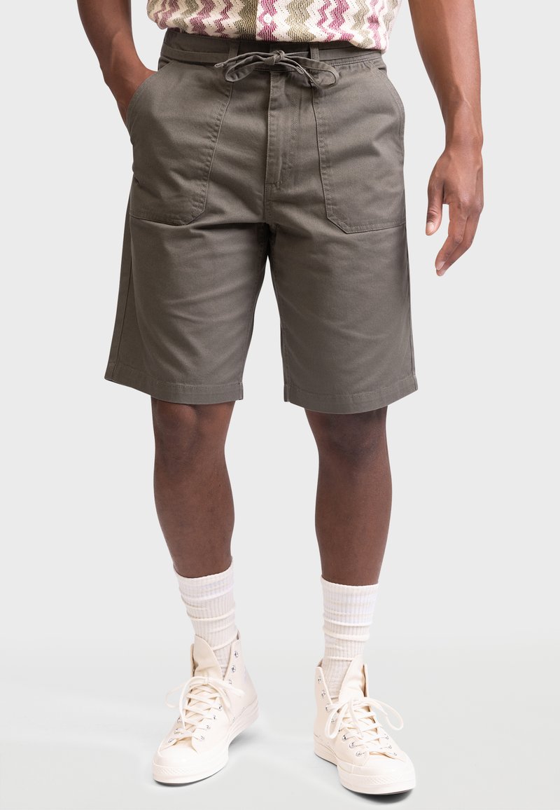 Pantaloni corti verde oliva con vestibilità rilassata, vita elastica e tasche frontali. Indossati con sneakers bianche alte e calze a coste.