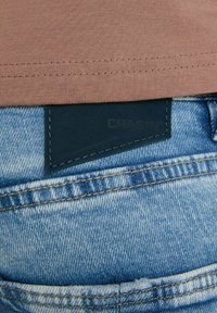 Una camicia di cotone marrone è sovrapposta a jeans di denim blu. Un'etichetta in pelle scura con la dicitura "CHASIN'" è visibile sui jeans, con cuciture a contrasto.