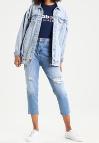 Džínová bunda se světlým praním, zapínání na knoflíky a dvěma náprsními kapsami, kombinovaná s džínovými kraťasy světle modré barvy s opotřebením a bílými teniskami.