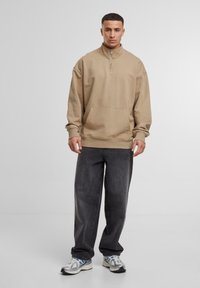 Beige zip-up hoodie met een buidelzak, gecombineerd met losse donkergrijze jeans en zilveren sportschoenen, staand tegen een lichte achtergrond.