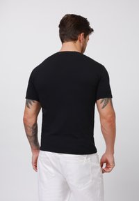 Black cotton t-shirt with a crew neckline, short sleeves, and a fitted shape, worn with white pants, showing a tattoo on the arm.