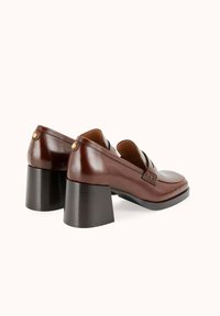 Mocassins en cuir marron avec un embout carré, deux brides et un talon épais noir. Présente une finition texturée et des accents dorés subtils.