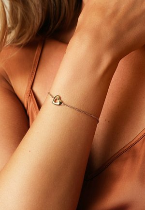 Delicato braccialetto con catena d'oro e piccolo ciondolo a cuore traforato, indossato al polso di una donna con top marrone senza maniche.