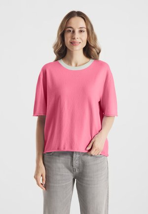 Vrouw met golvend lichtbruin haar, gekleed in een losse roze top met korte mouwen, een witte kraag en lichtgrijze broek, staand voor een witte achtergrond.