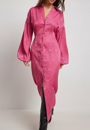 Vrouw draagt een roze satijnen blousejurk met lange mouwen, subtiele patronen en zwarte kniehoge laarzen, staand tegen een effen achtergrond.