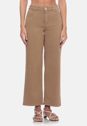 Donna che indossa pantaloni khaki a vita alta e gamba larga e sandali bianchi con tacco e cinturini, in piedi davanti a uno sfondo semplice.