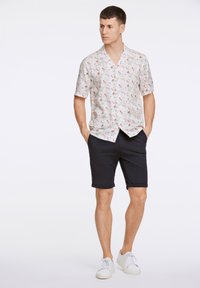 Camisa de manga corta con estampado floral en blanco y patrones coloridos; combinada con pantalones cortos negros y zapatillas blancas, mostrando un atuendo casual.