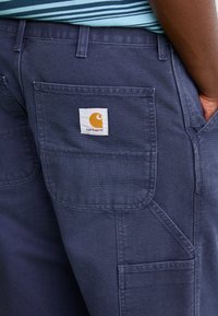 Navy work pants with multi pockets, reinforced stitching, and a white tag featuring a yellow logo. Cotton material with a durable texture.