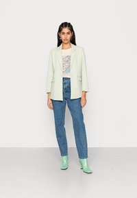 Blazer verde claro, de mangas curtas, combinado com uma t-shirt creme com gráfico e jeans azul de cintura alta. Botas verdes até os tornozelos completam o look.