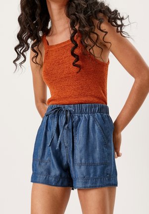 Mujer con top de punto sin mangas color óxido y pantalones cortos de mezclilla azul de cintura alta con cordón y bolsillos delanteros.