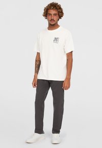 Hvid bomulds-t-shirt med kortærmer og et grafisk print på venstre bryst, parret med mørkegrå bukser og hvide sneakers.