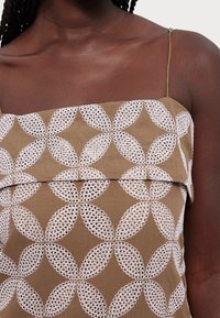 Brown fabric top featuring white floral lace patterns with overlapping circular designs. Thin adjustable straps and a layered look.