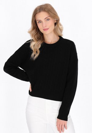 Schwarzer Kabelstrickpullover mit rundem Halsausschnitt und langen Ärmeln, mit einer lockeren Passform und geripptem Saum. Kombiniert mit weißen Hosen.