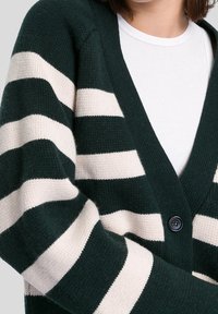 Vihreä ja kerma raidoitettu neuletakki, jossa on V-pääntie, etunapit ja teksturoitu neulemateriaali. Lähikuva hihasta ja päällekkäin olevasta kankaasta.