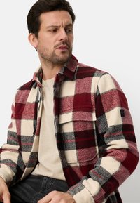 Rød og creme ternet flanneljakke med sorte detaljer, to brystlommer og en krave, båret over en beige sweater. Blød tekstur.