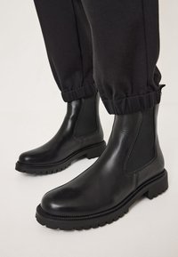 Botas negras de cuero hasta el tobillo con paneles laterales elásticos, suela de goma texturizada y punta redonda. El diseño presenta un acabado suave y costuras sutiles.