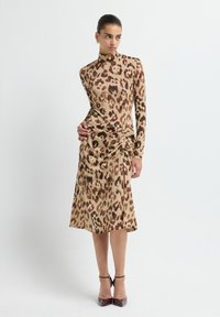 Donna in posa con un vestito a maniche lunghe, collo alto, al ginocchio, con stampa leopardata marrone, indossando tacchi neri con cinturino alla caviglia e orecchini dorati.