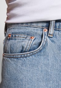 Jeans de denim azul claro com um pequeno bolso frontal, costura laranja e acentos de metal cobre. A textura aparenta ser suave com um design clássico.