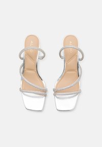 Sølvhælede sandaler med en glansfuld overflade, firkantet tå og flere sammenflettede remme med metalliske detaljer. Lysebrune indlægssåler.