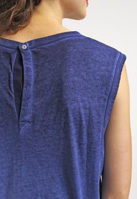 Kortärmad blå topp i mjukt tyg, med en nyckelhålsöppning på baksidan med en knappdetalj och en något avslappnad passform.