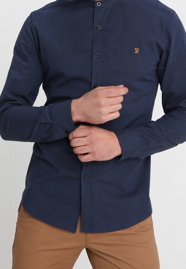 Man die de manchet van een nauwsluitend donkerblauw overhemd met een klein oranje logo aanpast, gekleed in lichtbruine broek, tegen een effen achtergrond.