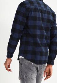 Chemise en flanelle à carreaux bleu marine et noir, à manches longues, avec des poignets boutonnés et une coupe droite. Portée avec un pantalon cargo gris.