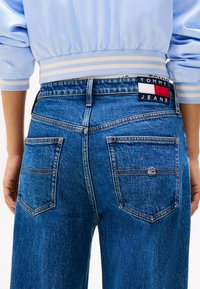 Bakre utsikt av person som har på seg blå Tommy Jeans denim med logo-lapp over høyre baklomme og lyseblå jakke med stripete mansjetter.