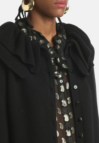 Schwarze Strickjacke mit rüschigem Kragen, mit Knopfverschluss. Über einer durchsichtigen Bluse mit goldenen Punkte-Mustern getragen.