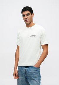 Biała bawełniana koszulka z okrągłym dekoltem, krótkimi rękawami i małym czarnym logo nadrukowanym na lewej piersi. W połączeniu z niebieskimi dżinsami.