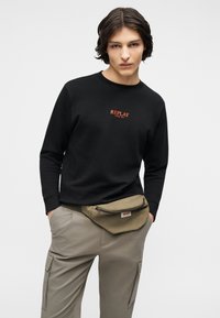 Νέος άνδρας με μακριά σκουρόχρωμα μαλλιά μέχρι τους ώμους, που φοράει μαύρο φούτερ "Replay", μπεζ παντελόνι cargo και χακί τσάντα μέσης.