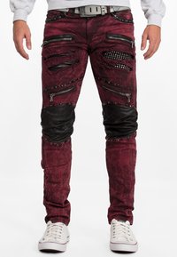 Jean bourgogne avec zones déchirées, accents en cuir, plusieurs poches zippées et détails cloutés ; style ajusté avec un subtil éclat.