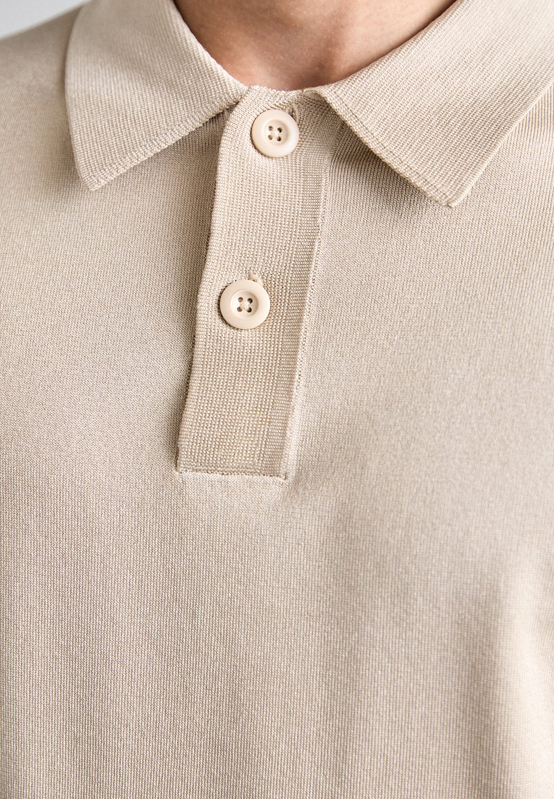 Beige pikétröja med en strukturerad stickning, klassisk krage och treknappsstängning med matchande knappar. Mjuk tygfinish.