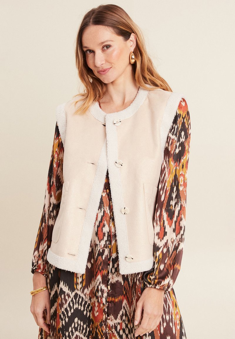 Beige Weste mit einem Lammfellbesatz, ausgestattet mit fünf runden Knöpfen und Seitentaschen. Das Modell trägt ein gemustertes Kleid darunter.