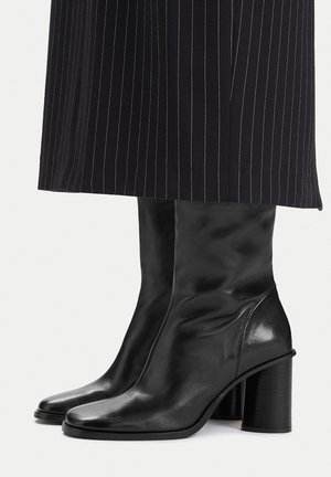 Botas negras de cuero hasta la mitad de la pantorrilla con tacones anchos usadas debajo de pantalones anchos negros con rayas diplomáticas.