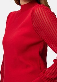 Czerwona sweter z wysokim dekoltem i przezroczystymi, prążkowanymi rękawami. Główna część jest gładka w dotyku i dopasowana.