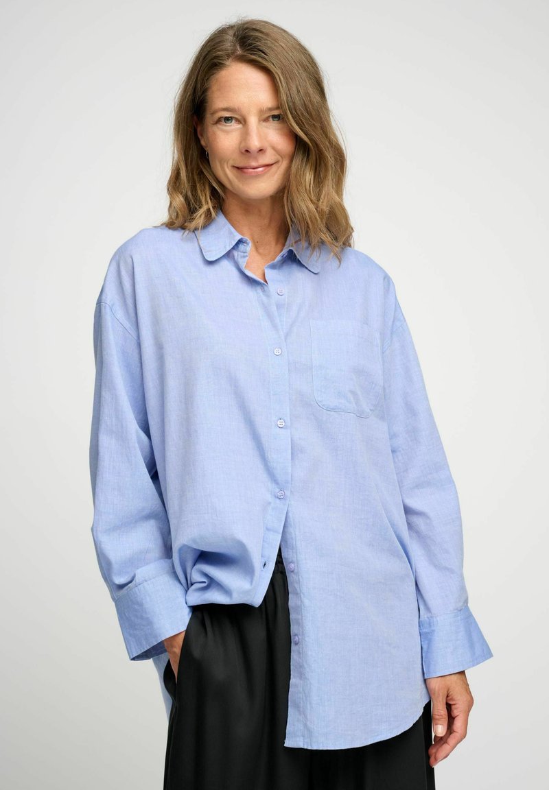 Camicia azzurra chiara con bottoni, realizzata in tessuto morbido. Presenta una tasca sul petto, colletto e maniche larghe. Abbinata a un pantalone nero.