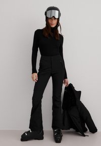 Kvinne iført svart skihjelm, skibriller, svart langermet overdel, skibukser, støvler, holder en svart vinterjakke mot en lys grå vegg.