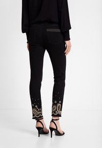 Mujer vistiendo jeans negros ajustados con bordado de paisley dorado en la parte baja de las piernas y una camiseta negra de mangas largas, combinada con sandalias de tacón alto negras.