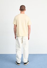 Beige bomulds T-shirt med korte ærmer, parret med hvide afslappede bukser og sorte sneakers med cremefarvede detaljer, set fra ryggen.