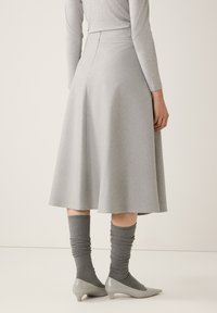 Frau trägt einen hellgrauen Midirock, ein passendes langärmliges Oberteil, graue gerippte Socken und hellgraue Schuhe mit kleinem Absatz, steht vor einem einfarbigen Hintergrund.