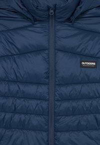 Veste matelassée bleu marine avec zip à l'avant et patch "OUTDOORS Expedition" sur le côté droit de la poitrine.