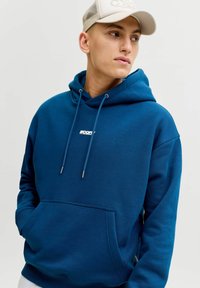 Jonge man met een blauwe hoodie met voorzak en trekkoordjes, en een beige baseballpet met gaas, kijkt naar rechts.