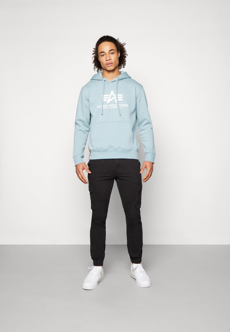 Lichtblauwe hoodie met een grafisch ontwerp aan de voorkant, verstelbare trekkoord en geribbelde zoom. Gecombineerd met zwarte cargobroek en witte sneakers.