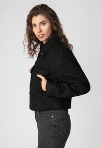 Black cropped jacket with a textured fabric, wide collar, and front pockets. Paired with dark grey slim-fit jeans.