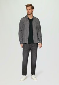 Szary koszulowy shirt w drobne paski, czarna koszulka z dekoltem w serek pod spodem, ciemne dżinsy i białe sneakersy. Model stoi na jednolitym tle.